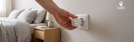 Une main branche un testeur de prise dans une prise murale Schuko à côté d’un lit, ses témoins allumés