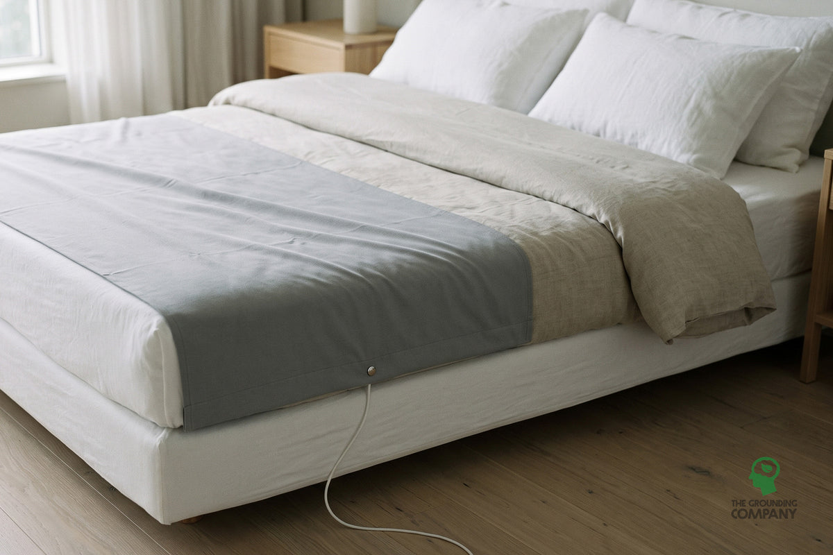 A grey grounding half sheet in use across the lower half of a made bed, with its connection cord and snap stud visible running down to the floor.