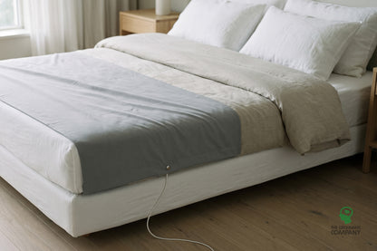 A grey grounding half sheet in use across the lower half of a made bed, with its connection cord and snap stud visible running down to the floor.